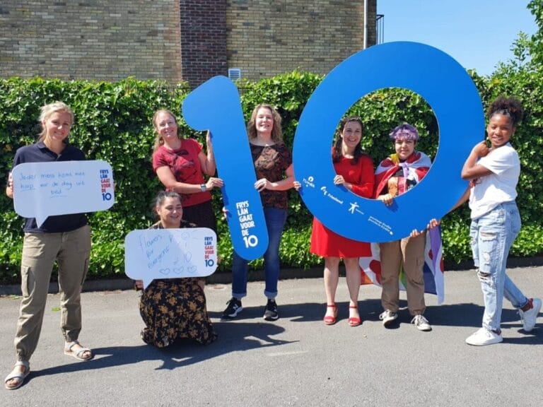 Groepsfoto van collega's in het project 'Fryslân gaat voor de 10'. Zeven menen staan naast elkaar op de foto en houden een grote blauwe 10 vast.
