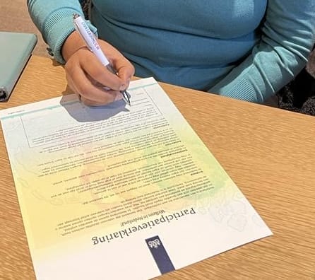 Iemand houdt een pen vast en ondertekent de Participatieverklaring.