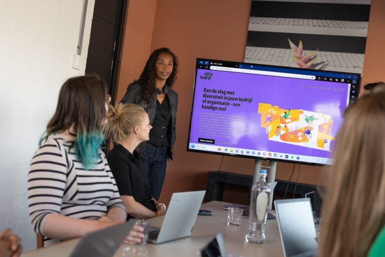 Een trainer staat voor de groep en geeft een presentatie. Op de presentatie zien we de tekst: 'Aan de slag met diversiteit in jouw bedrijf of organisatie - een handige webtool'.
