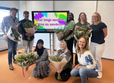Op de foto staat een groep mensen die meegewerkt hebben aan de korte film 'Over lange mensen, gras en meedoen'. Zij staan op de foto met bossen bloemen, omdat zij bedankt zijn voor hun bijdrage aan de film. Achter hen staat een display met daarop de titel van de film: 'Over lange mensen, gras en meedoen'.