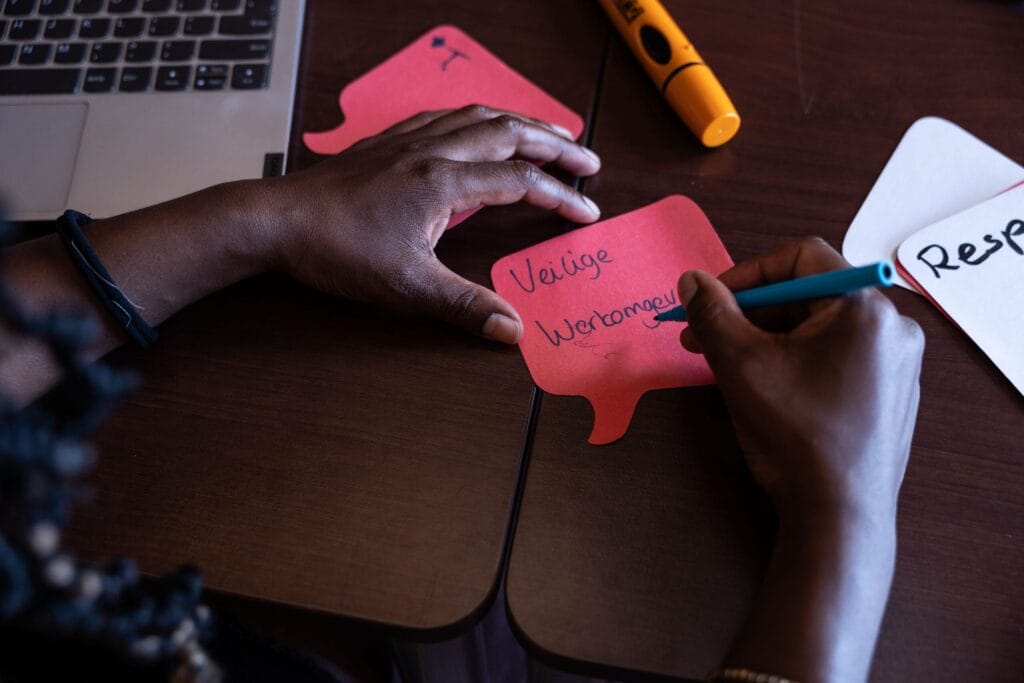Op de foto staan twee handen afgebeeld. De persoon houdt een pen vast en schrijft op een rode post it 'veilige werkomgeving'
