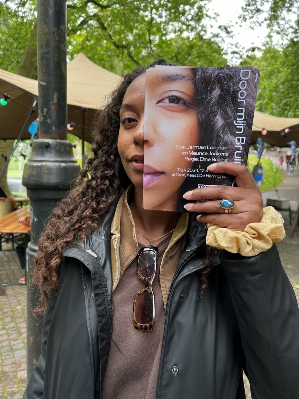 Collega Marem Kane die een flyer van de theatervoorstelling 'Door Mijn Bruine Ogen' voor haar gezicht houdt. Leer om te gaan met racistische kreten zoals "Ga terug naar je eigen land" door middel van deze voorstelling.