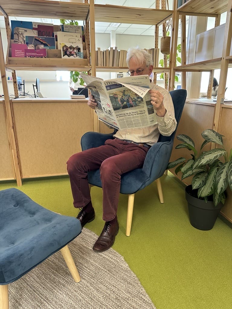 Een foto van Collega Harry Prins die de Leeuwarder Courant vast heeft. Hij bevindt zich op het kantoor van Tûmba en zit op een blauwe stoel voor een houten kast.