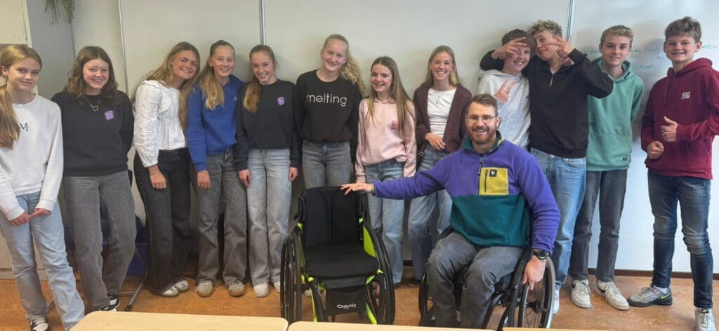 Een groepsfoto gemaakt na een voorlichting op het middelbaar onderwijs. Hierop zie je kinderen staan en voor de kinderen zit Rienk in een rolstoel en naast Rienk staat een sportrolstoel in de kleur neongeel.